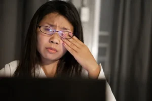 Woman wearing glasses sits at a laptop, rubs her eye in apparent fatigue or discomfort.